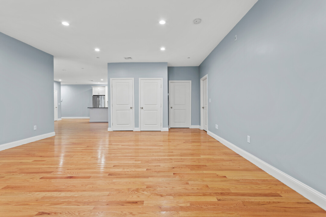 Empty room with hardwood floors and pale blue walls, facing double closet doors with the kitchen visible to the left.