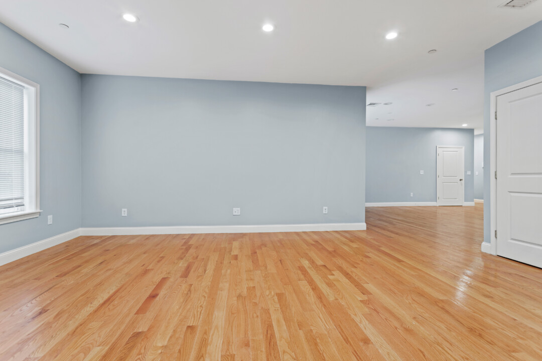 Empty living area with hardwood floors, pale blue walls, recessed lights, and an opening to another part of the apartment.