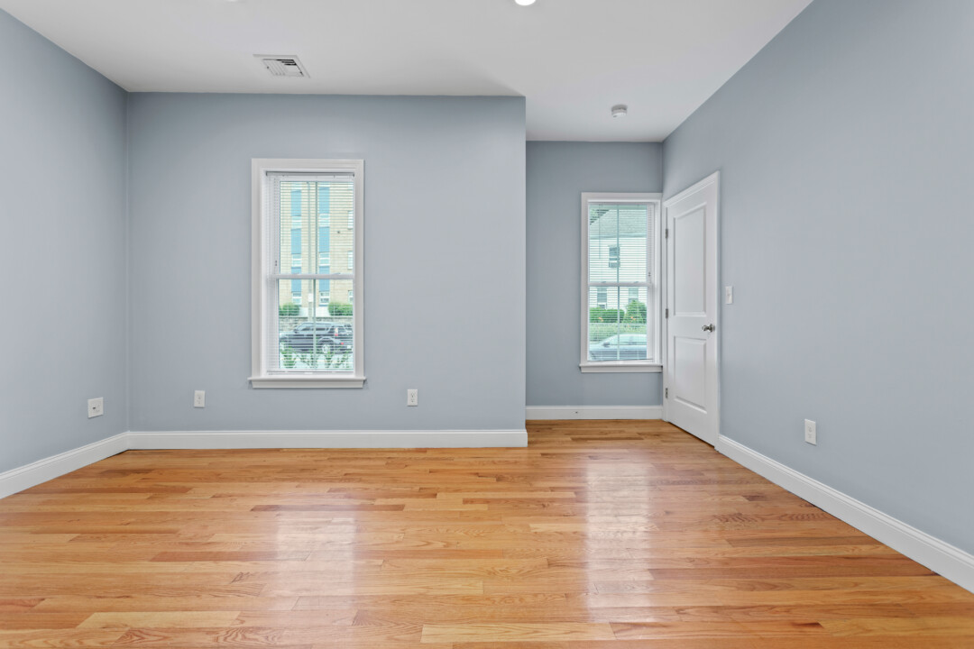 Empty bedroom with hardwood floors, pale blue walls, two windows, and a white door.