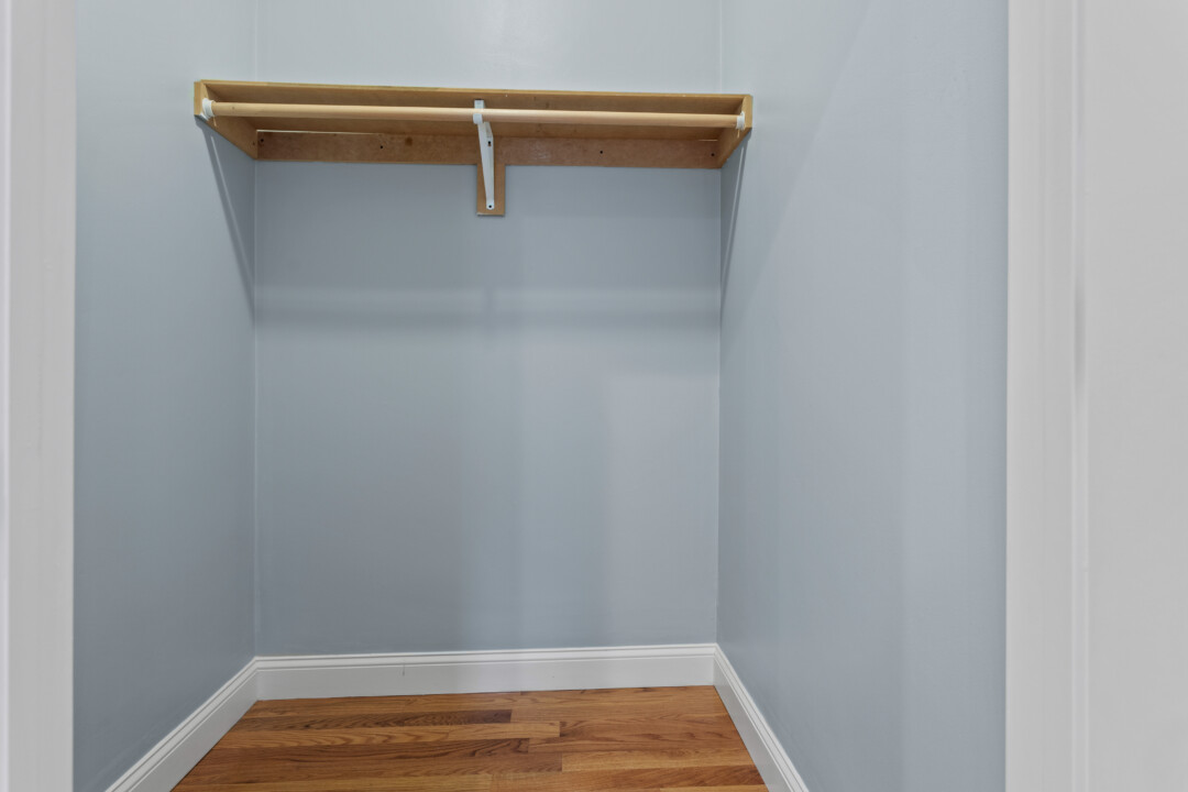Empty closet with pale blue walls, hardwood flooring, and a wooden shelf and hanging rod.