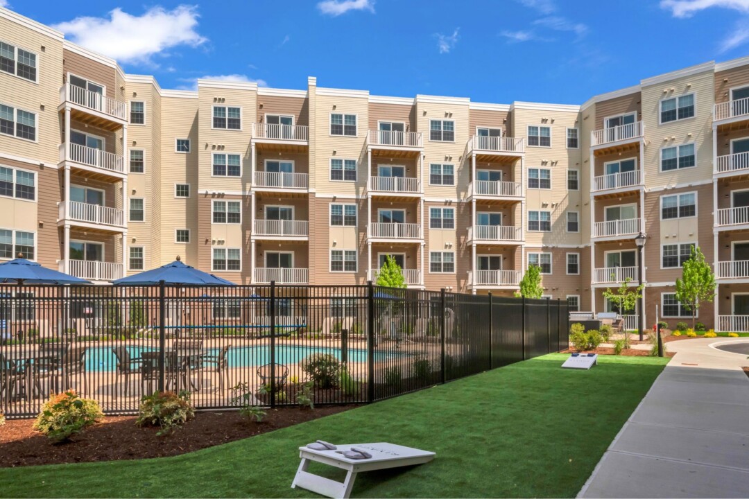 Grassy courtyard with a cornhole board beside a fenced pool. A multistory tan and brick apartment building with balconies surrounds the courtyard.