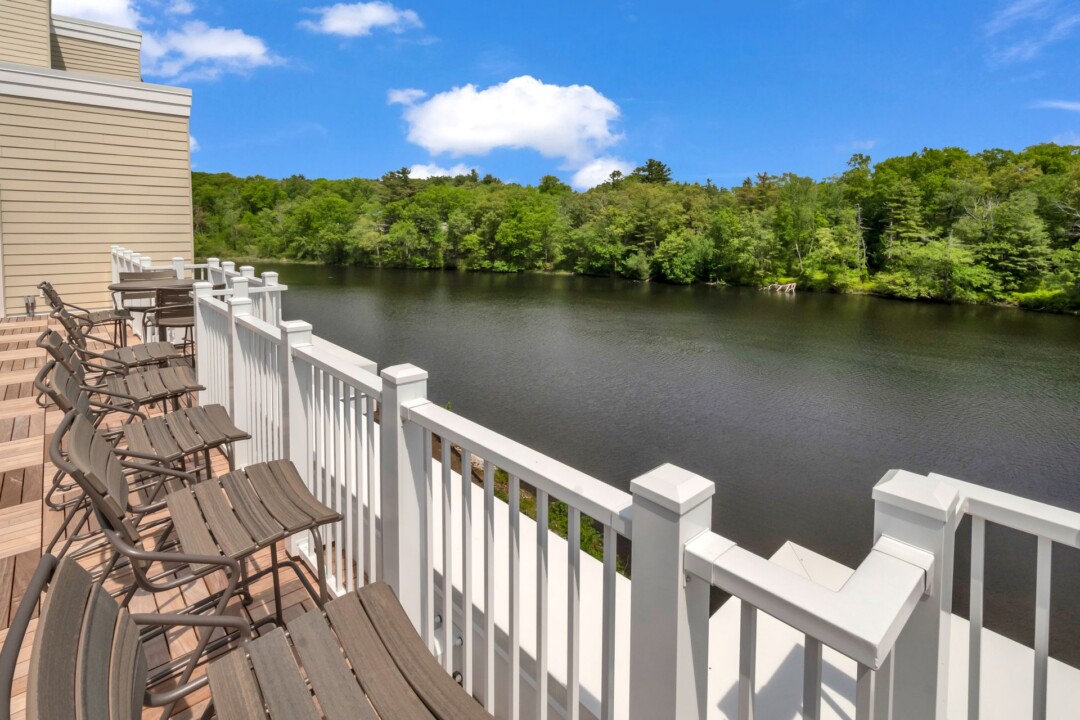 Wooden deck with chairs and a white railing overlooking a river and tree-lined shore.