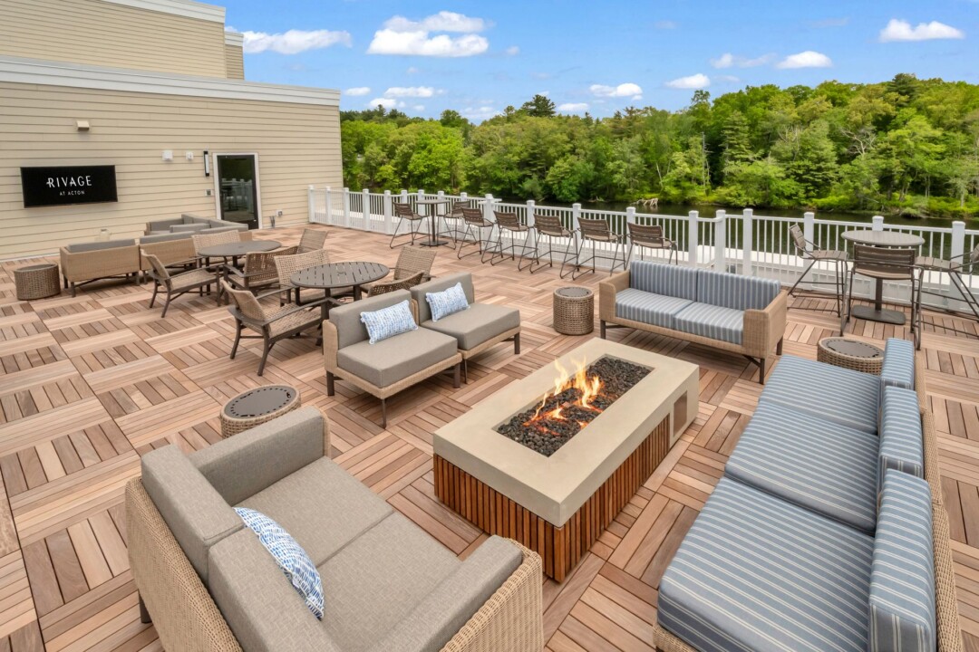 Rooftop terrace with cushioned sofas and chairs arranged around a rectangular fire pit, with a white railing and trees beyond.