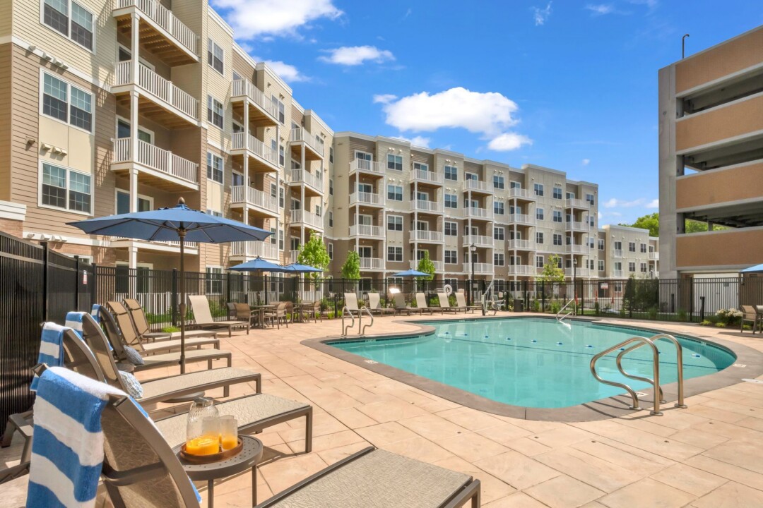 Outdoor pool beside a multistory apartment building with tan siding, brick accents, and balconies. Lounge chairs and blue umbrellas sit along the pool deck.