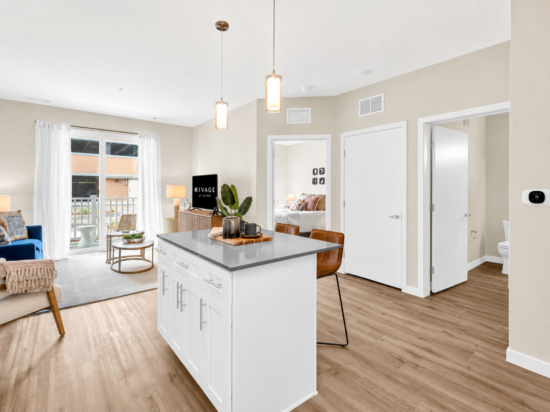 Open-plan apartment with a white kitchen island, pendant lights, dining space, and living area by large windows.