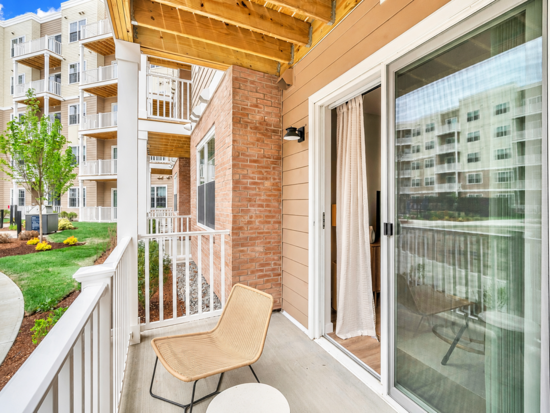 Apartment balcony with a chair, white railing, and sliding glass door. Brickwork and neighboring balconies are visible along the building.