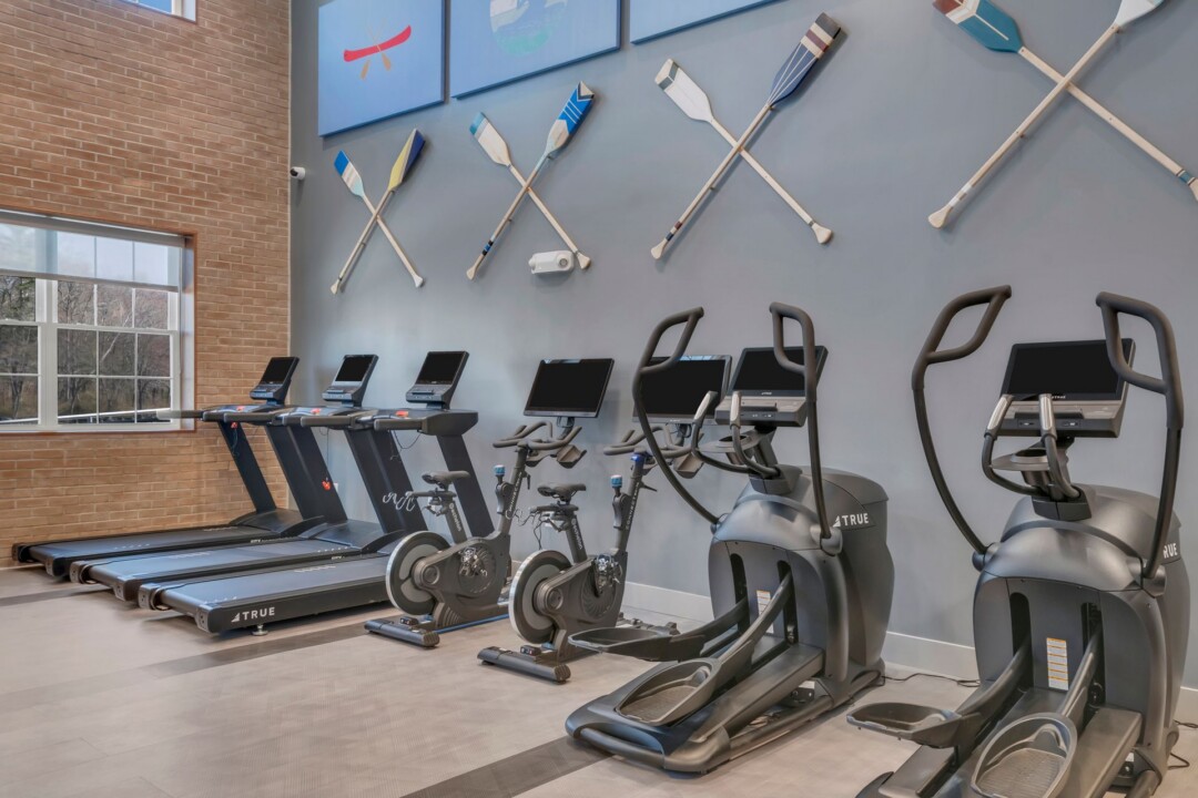 Fitness room with treadmills, stationary bikes, and elliptical machines beneath a wall display of sports equipment.