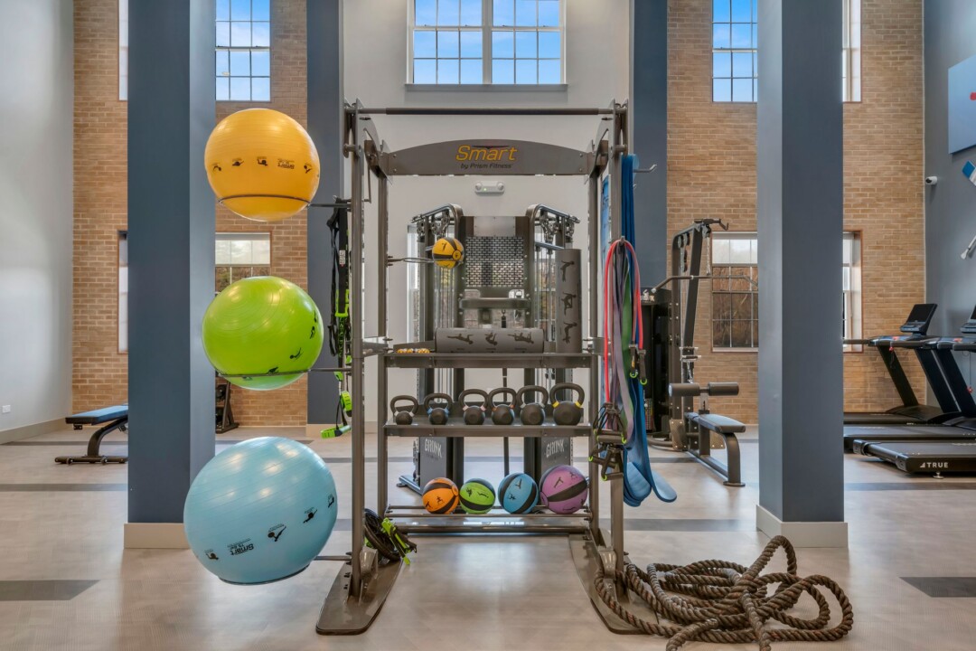 Fitness area with exercise balls, a weight rack, and strength-training equipment in front of tall windows.