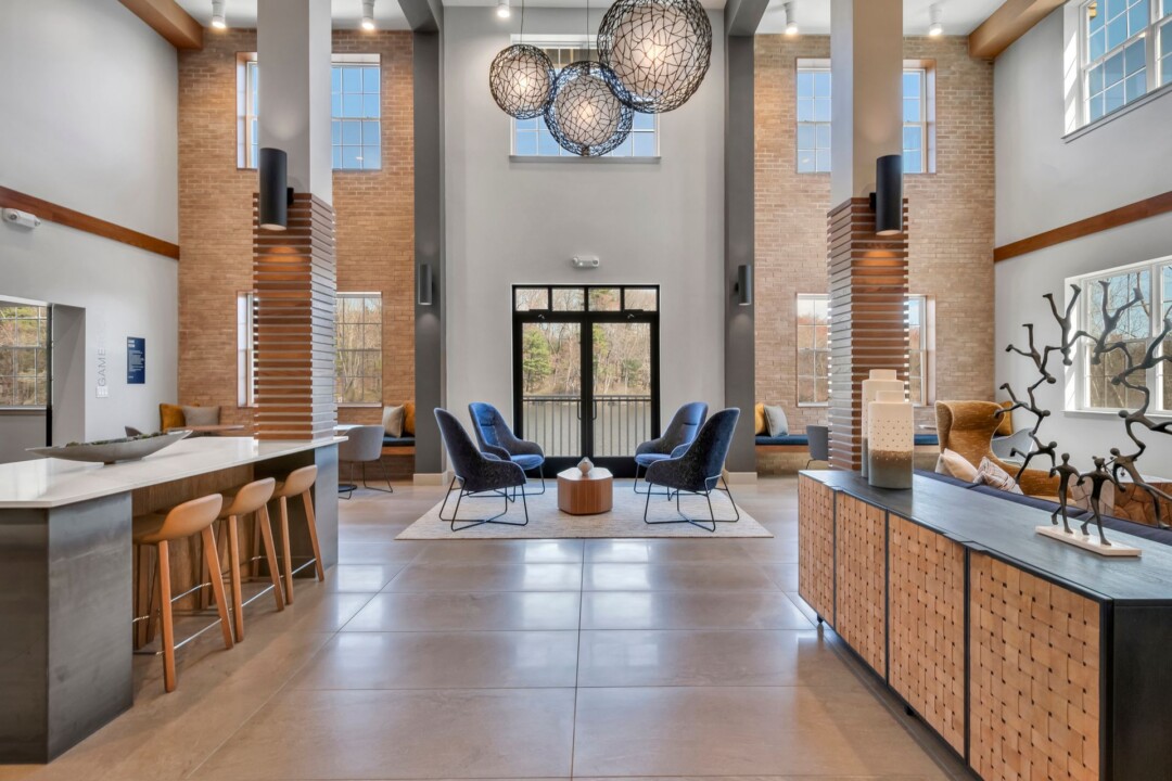 Apartment lobby with a high ceiling, brick columns, large windows, and blue chairs around a seating area.