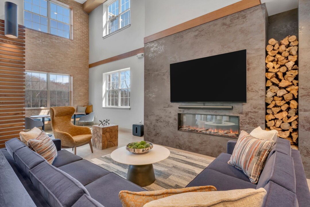 Resident lounge with sofas, armchairs, and a round coffee table facing a wall-mounted television above a fireplace. Brickwork and tall windows frame the room.