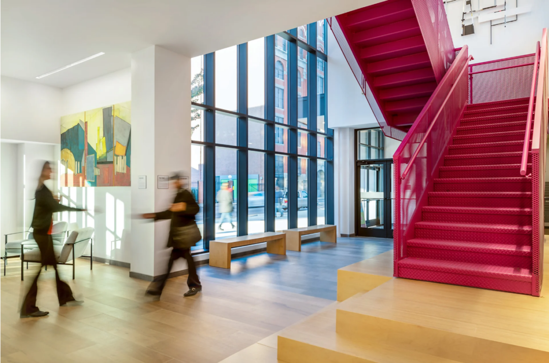 Lobby with a bright pink staircase, tall windows, wooden benches, and a seating area.
