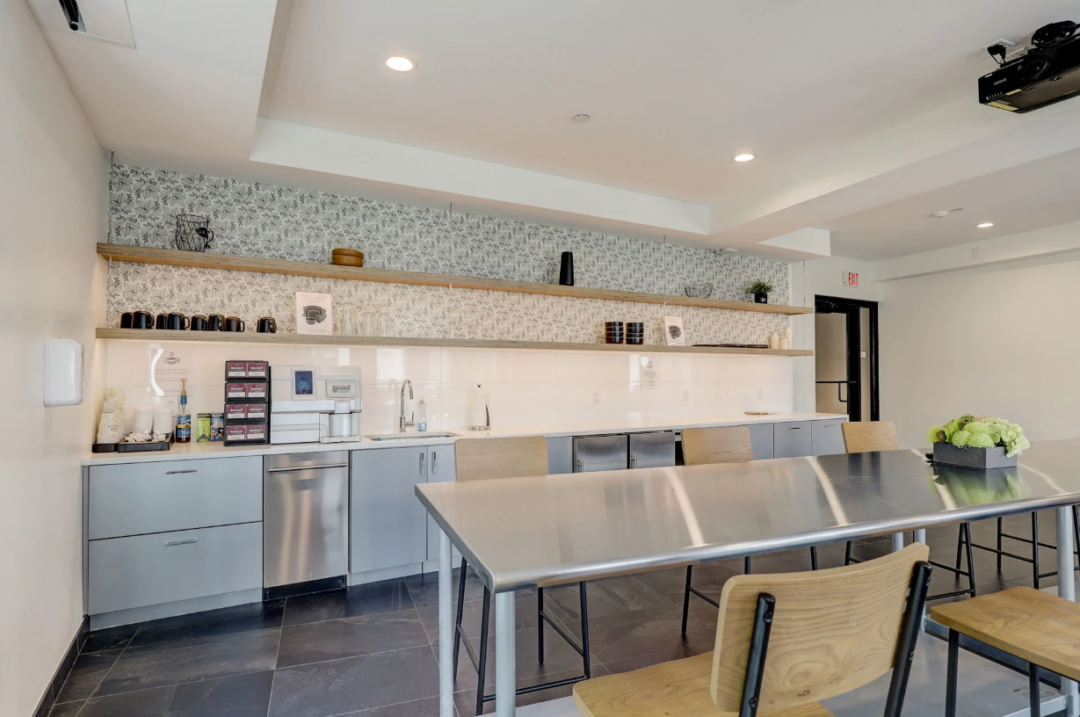 Shared room with a long table, stools, a kitchenette, and a ceiling-mounted projector.