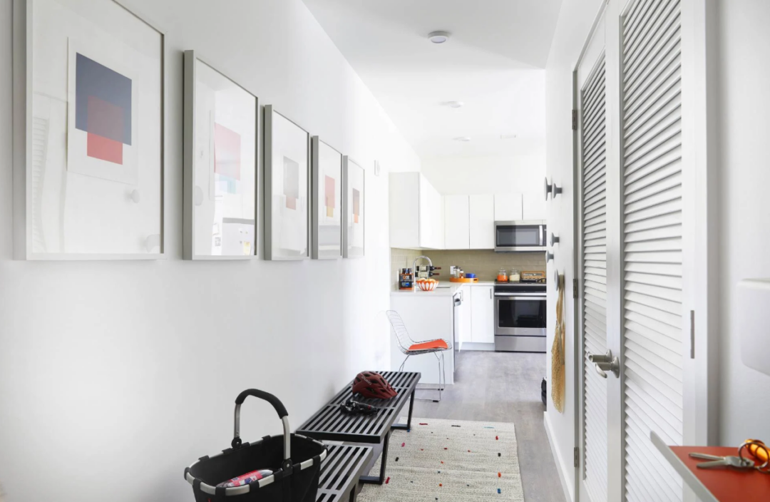 Hallway with framed artwork, benches, and louvered closet doors leading to a kitchen.