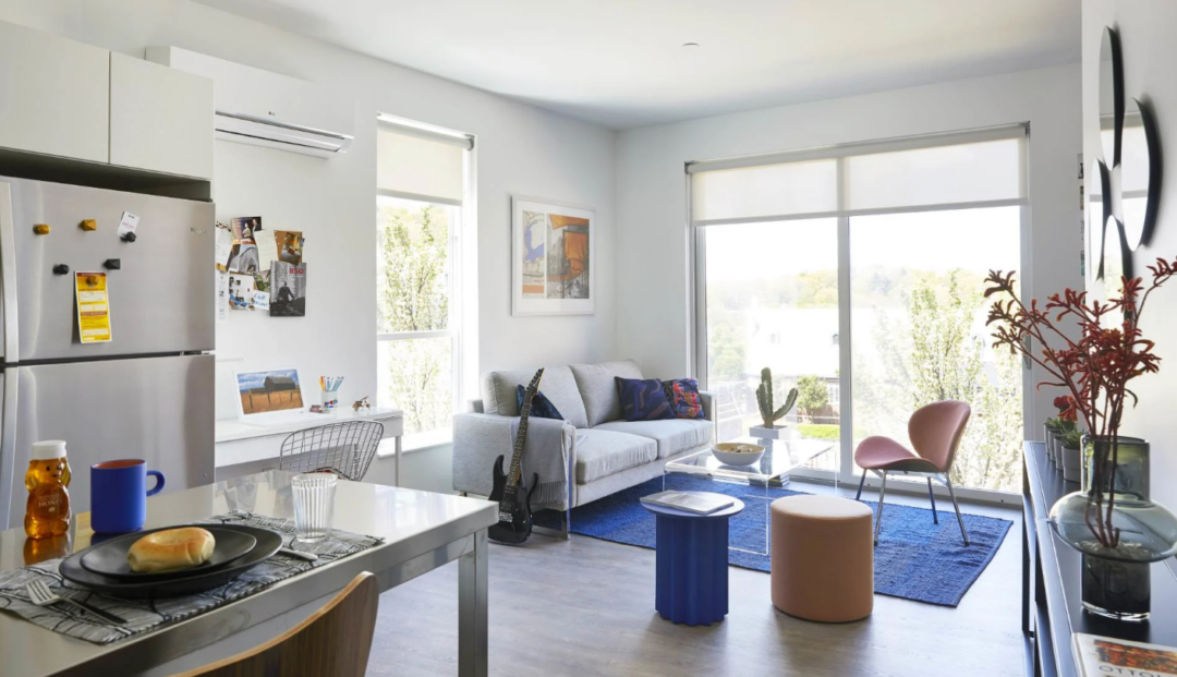 Open living area with a gray sofa, desk, pink chair, and large windows.