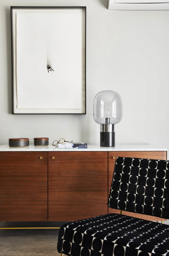 Patterned black chair beside a wooden cabinet with a glass lamp and framed wall art.