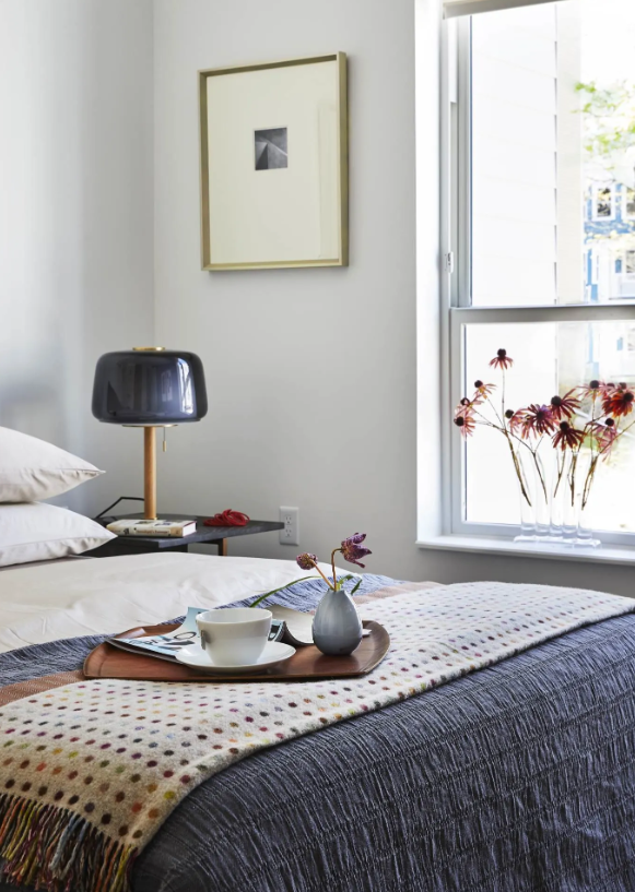 Bed with a breakfast tray beside a lamp and a window with flowers on the sill.