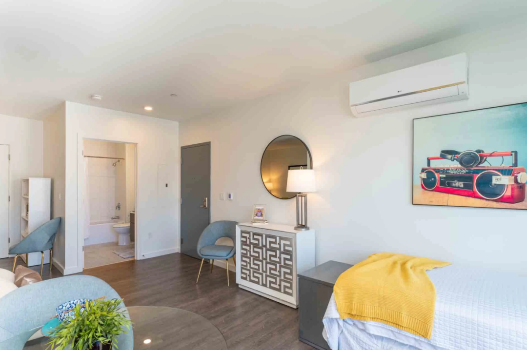 Furnished room with a bed, chairs, decorative cabinet, and an open bathroom doorway.
