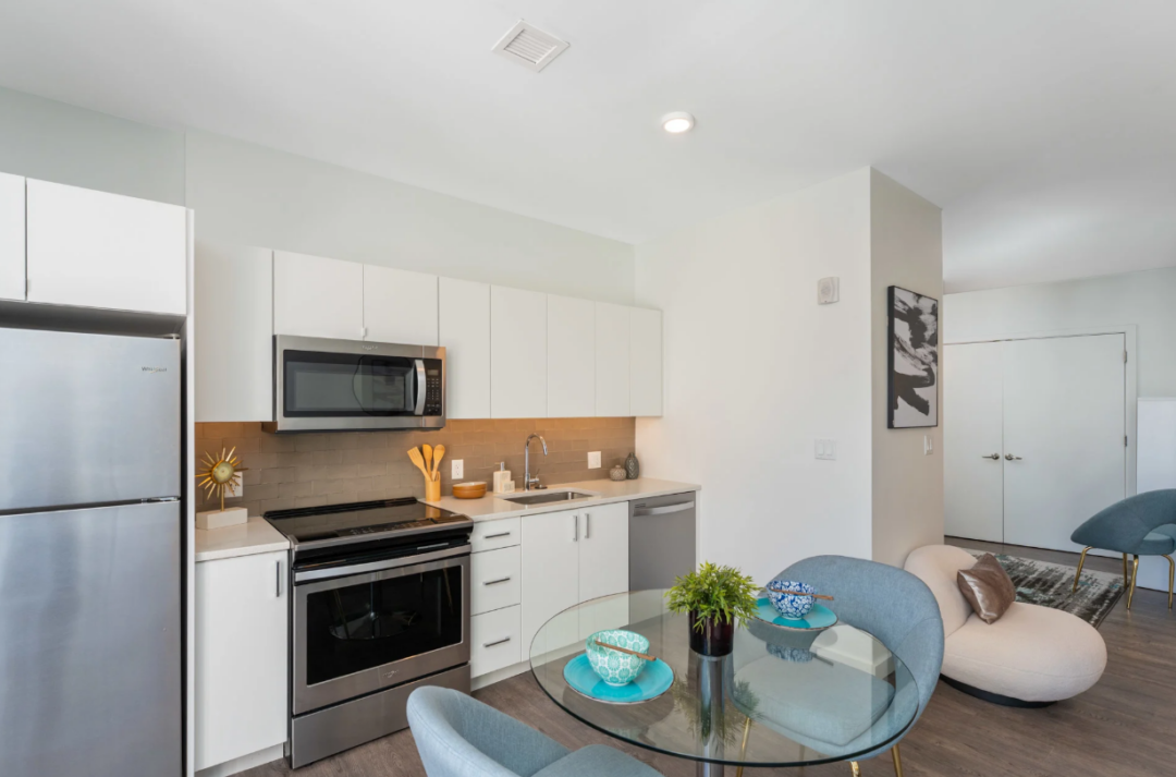 Kitchen with white cabinets and stainless steel appliances beside a glass dining table with blue chairs.