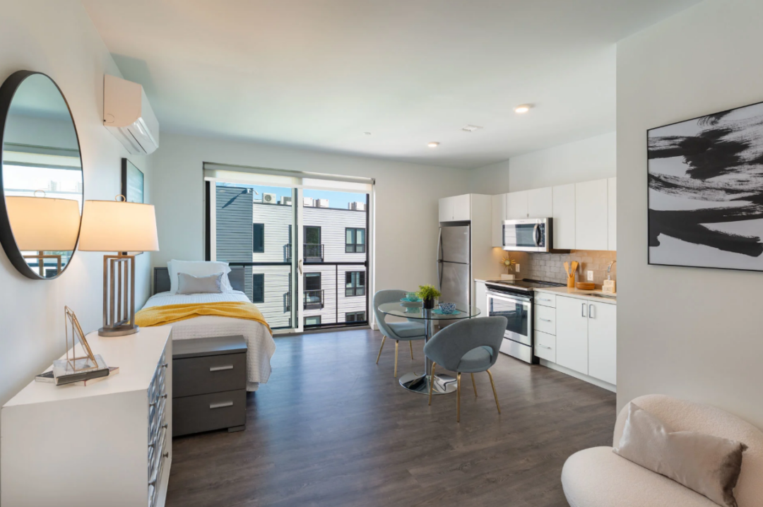 Studio apartment with a bed, glass dining table, kitchen, and large windows.
