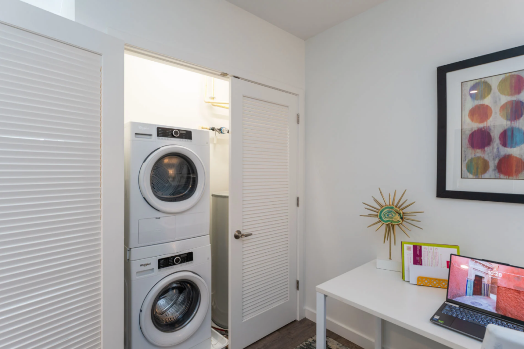 Stacked washer and dryer in an open closet beside a desk with a laptop.