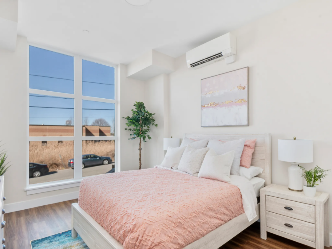 Bedroom with a pink bedspread, white furniture, a potted plant, and large windows overlooking nearby rooftops.
