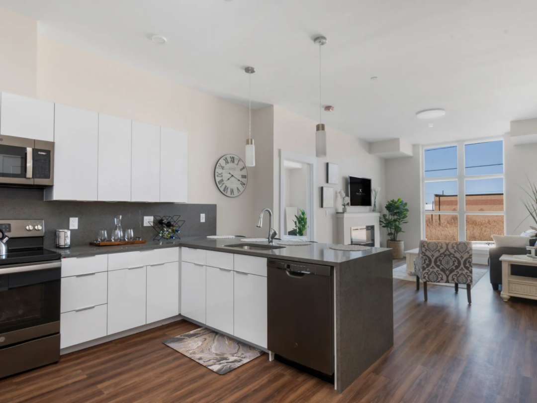 Another view of the kitchen with white cabinets, stainless steel appliances, wood-look flooring, and the adjoining living area.