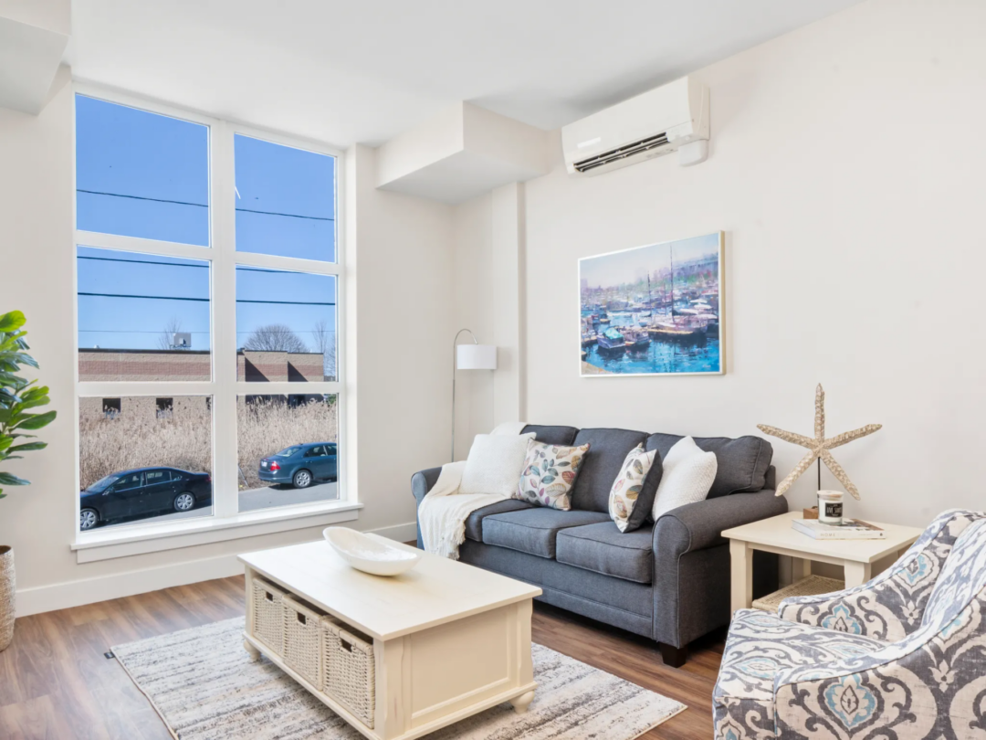 Living room with a dark gray sofa, wooden coffee table, patterned rug, and large windows overlooking nearby rooftops.