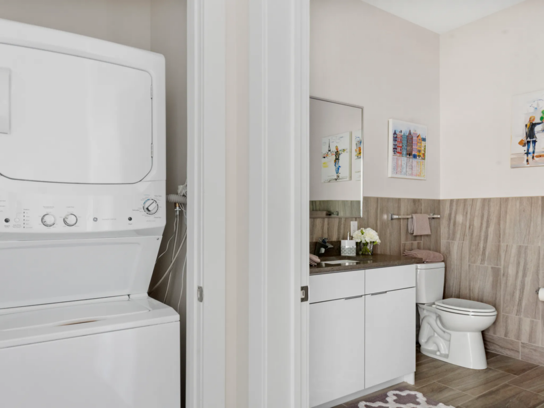 Laundry closet with a stacked washer and dryer beside a bathroom with a white vanity, mirror, and toilet.