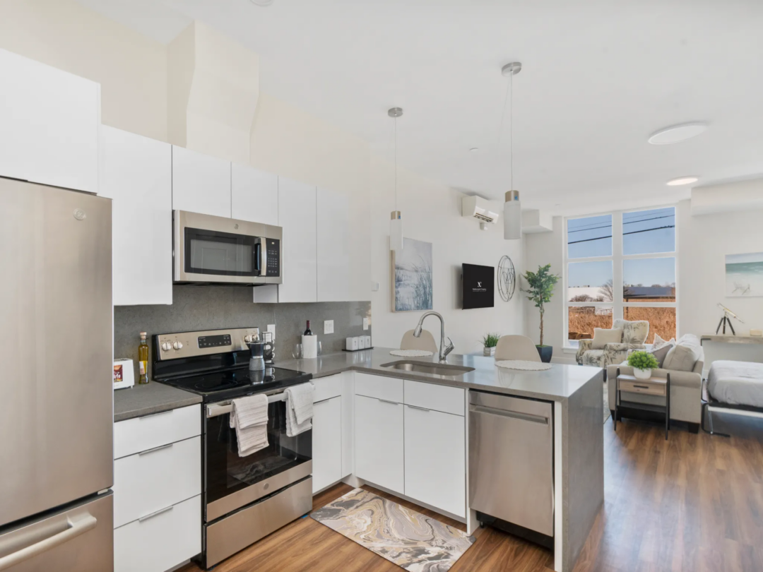 Apartment kitchen with white cabinets, stainless steel appliances, a gray backsplash, and a peninsula beside the living area.
