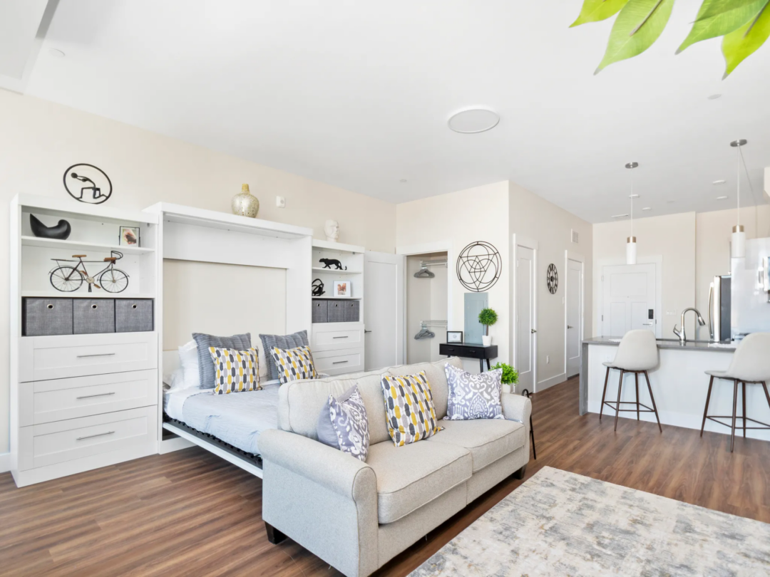 Studio apartment with a light gray sofa, patterned cushions, and an area rug, with a bed behind the sofa and a kitchen and breakfast bar to the right.