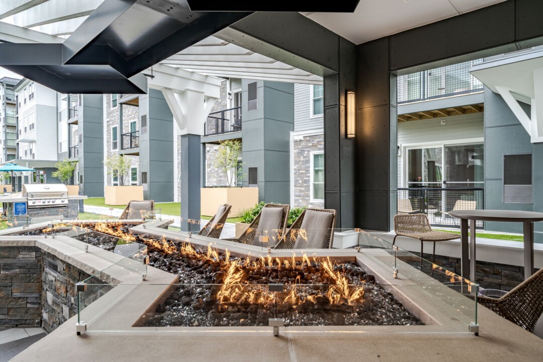 Covered outdoor lounge with a rectangular fire pit, seating, and views of the surrounding apartment buildings.