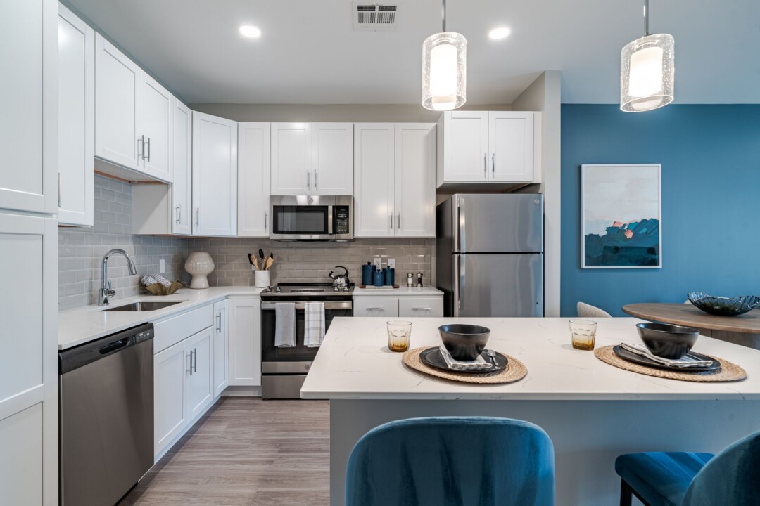 Wider view of the kitchen with white cabinets, stainless steel appliances, and an island with blue upholstered stools.