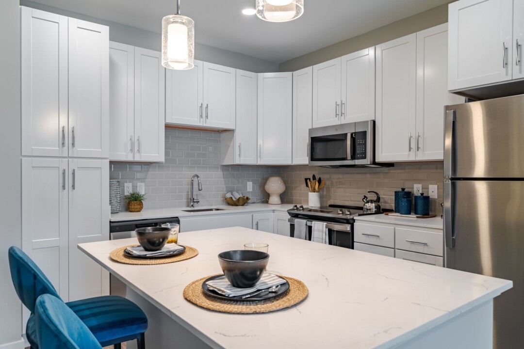 Apartment kitchen with white cabinets, stainless steel appliances, pendant lights, and a white island set with dishes.