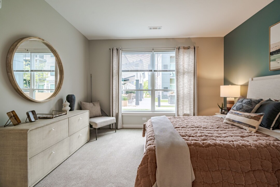 Another view of the bedroom with a dresser, round mirror, upholstered chair, and a large window beside the bed.
