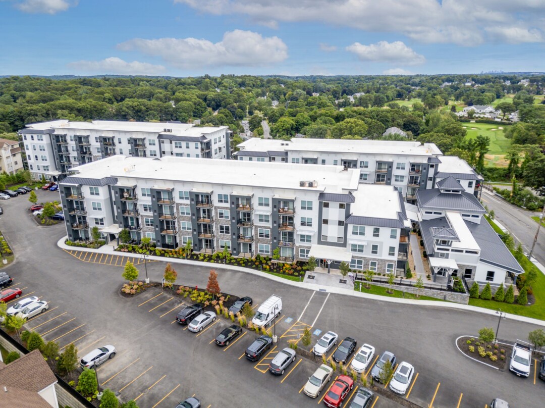Aerial view of a multistory apartment complex with white and gray exterior walls, balconies, and a parking lot, surrounded by trees.