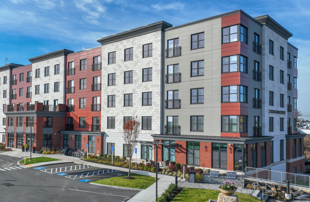Multistory apartment building with red, gray, and white exterior panels, rows of windows, landscaped grounds, and a parking area.